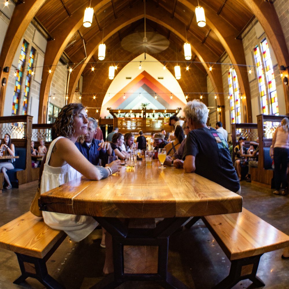 Woman seated at a long live-edge wood table inside a church-turned-taproom with arches and stained glass