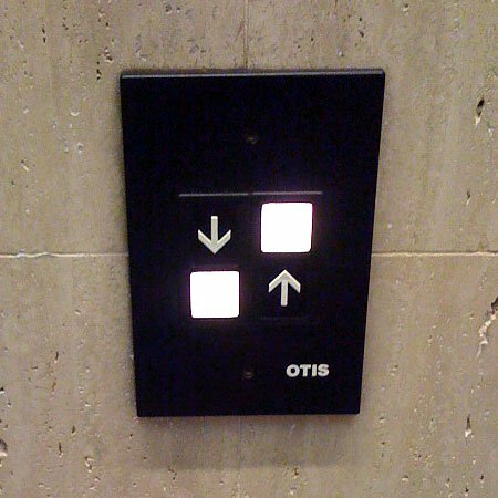 Black Otis elevator call panel with lit up and down arrow buttons on a stone wall