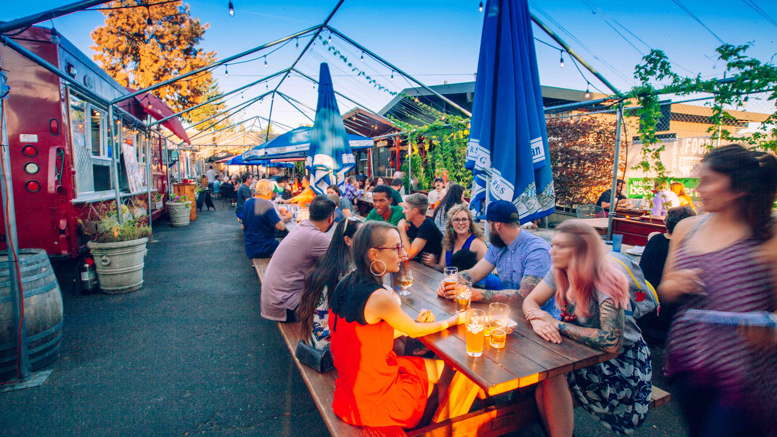 Evening crowd dining at long tables under string lights and umbrellas beside food trucks at Beergarden
