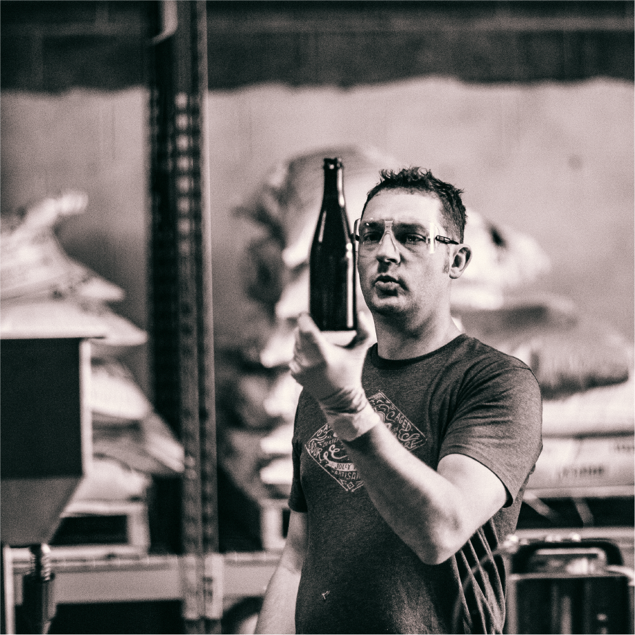 Black-and-white shot of a ColdFire brewer in safety glasses holding up a bottle to inspect it