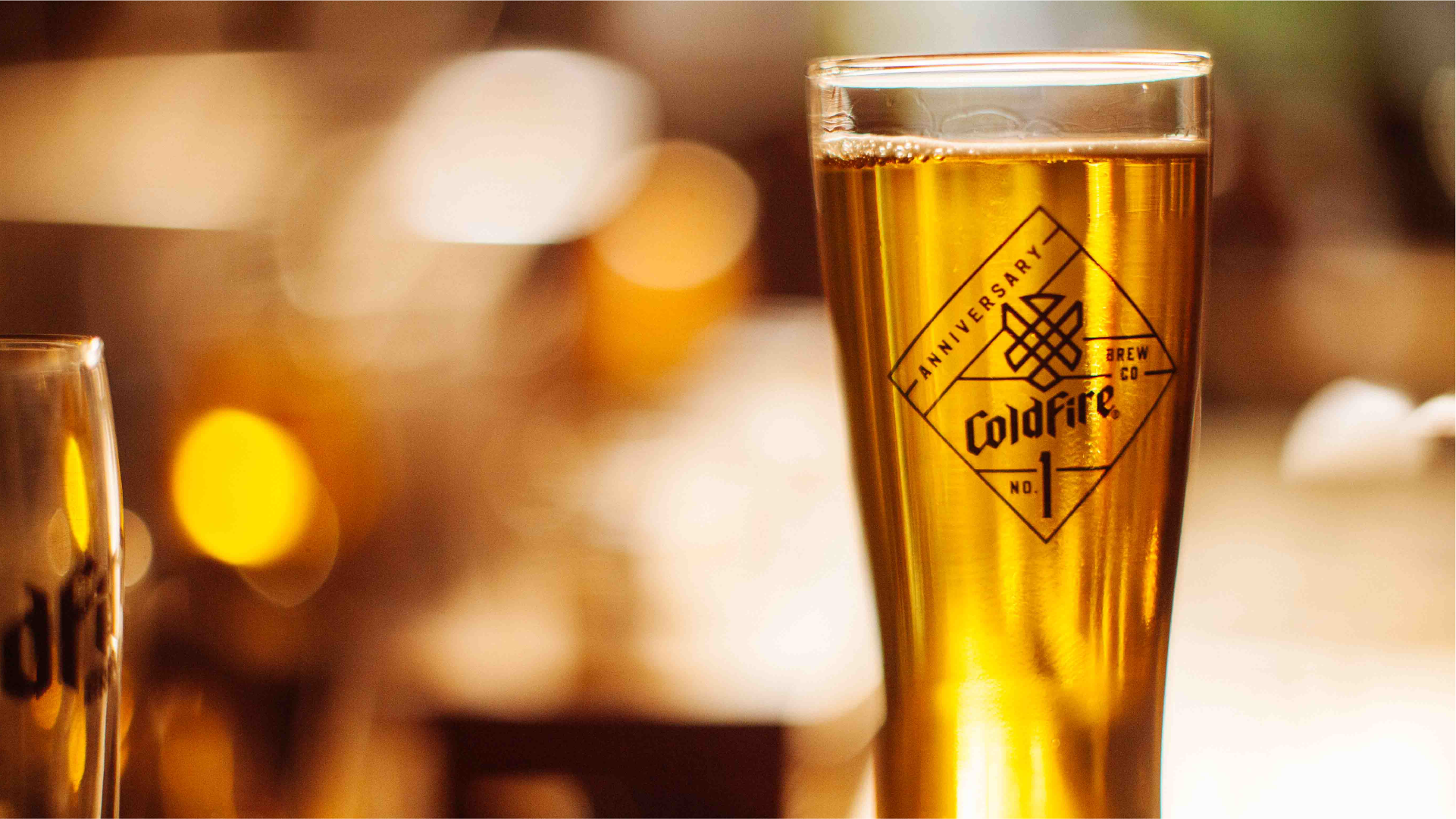 Tall glass of golden ColdFire beer with the anniversary diamond logo, warm bokeh lights behind