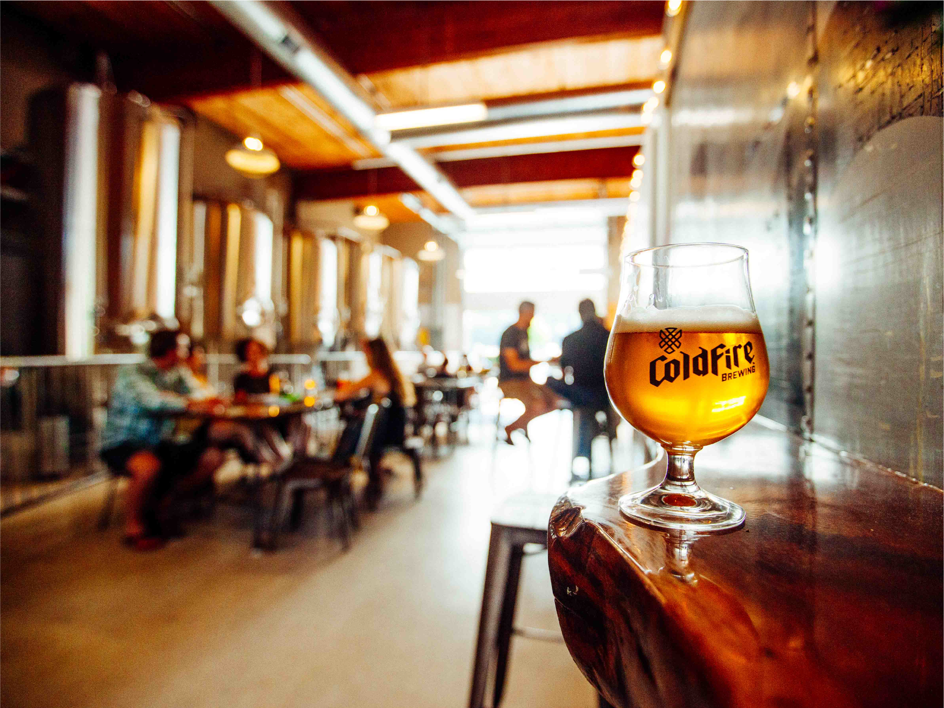 Glass of ColdFire beer on a wooden ledge with the brewery taproom and fermentation tanks blurred behind