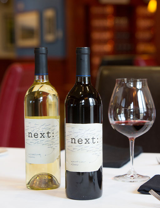 Two 'next:' wine bottles, white and red, with a glass of red wine on a restaurant table
