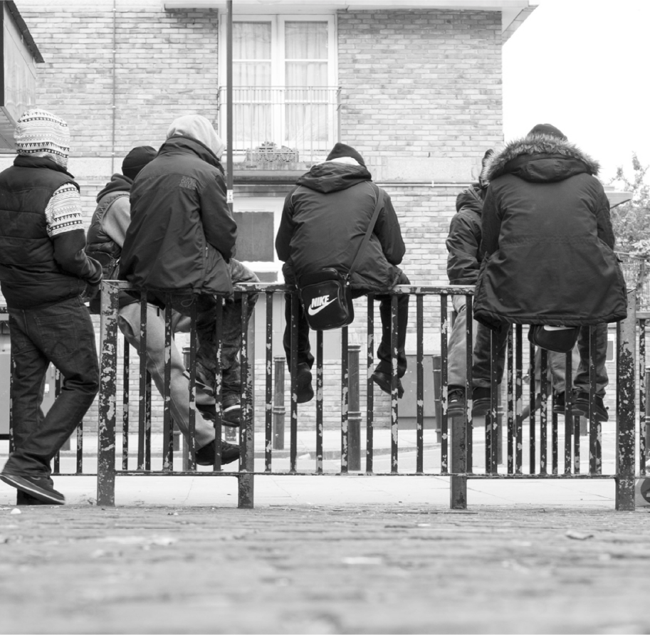 Young men in winter coats sit on metal railings outside a brick estate, one with a Nike bag, Nike London