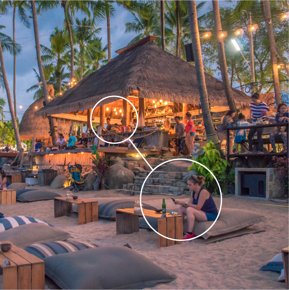 Thatched-roof beach bar at dusk with string lights, low tables and guests on cushions in the sand