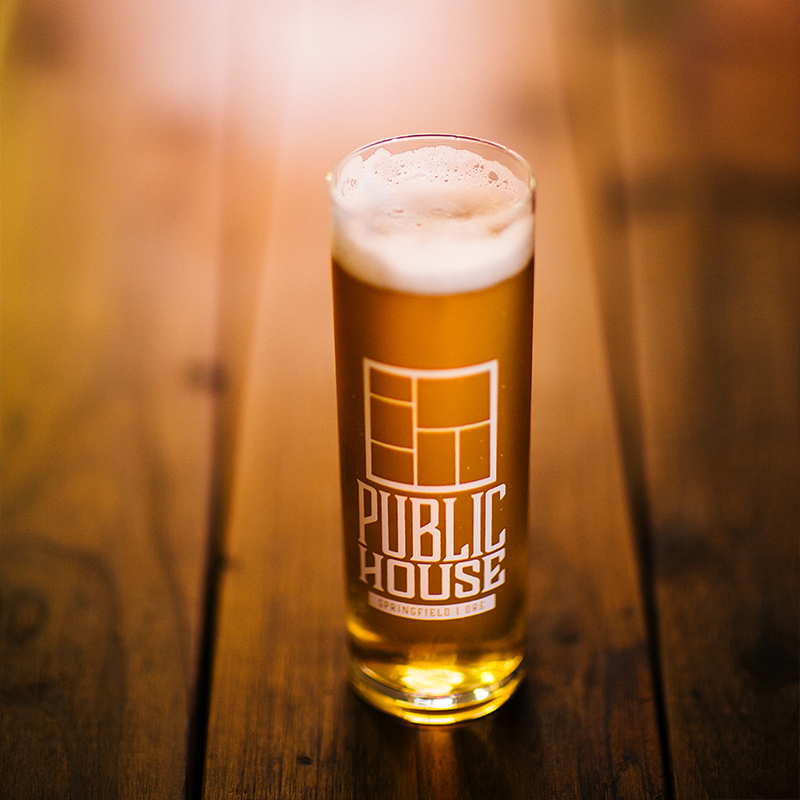 Glass of pale beer etched with the Public House Springfield Ore logo on a wood plank table
