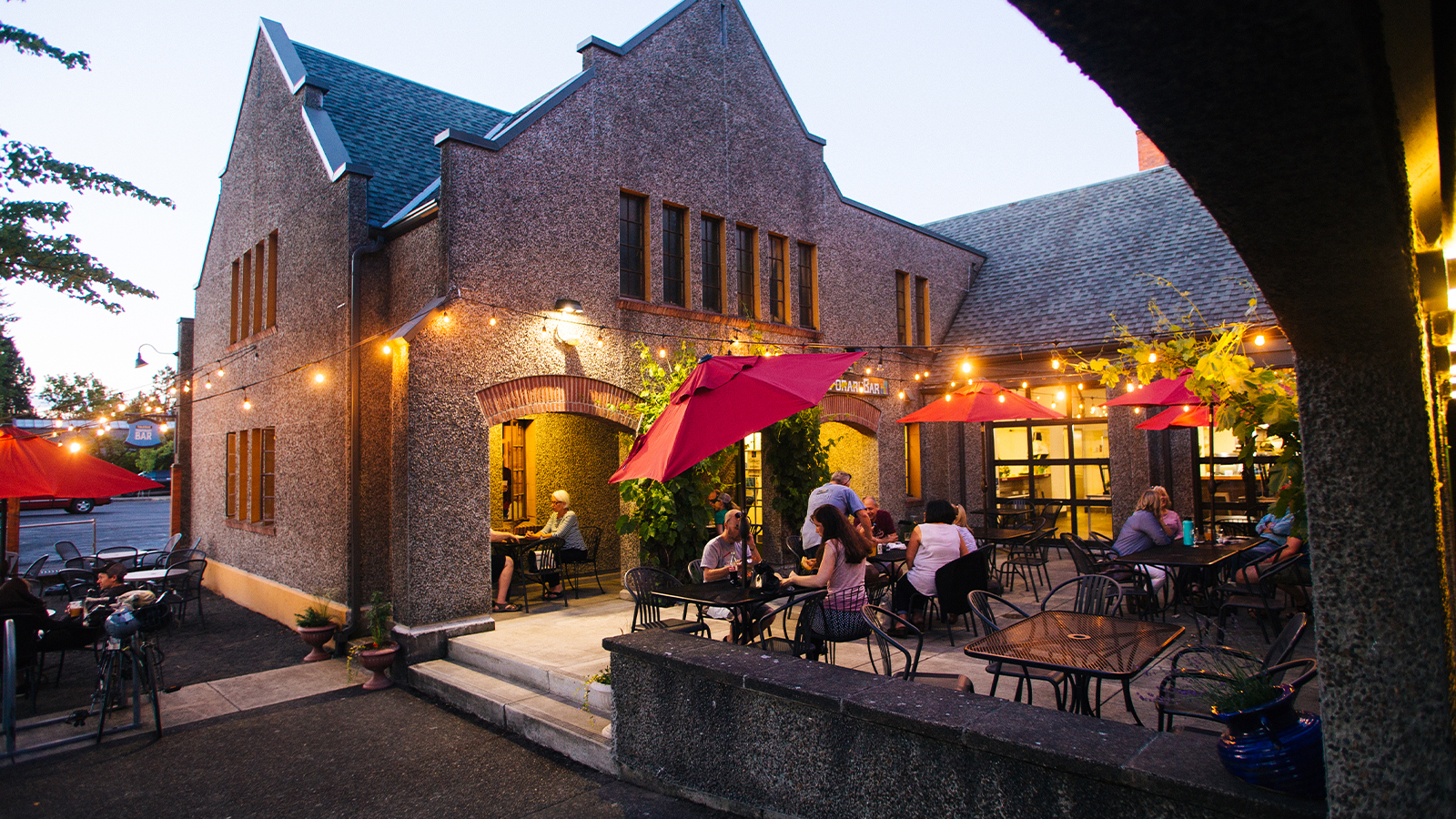 Stone Public House building at dusk with patio diners under red umbrellas and string lights