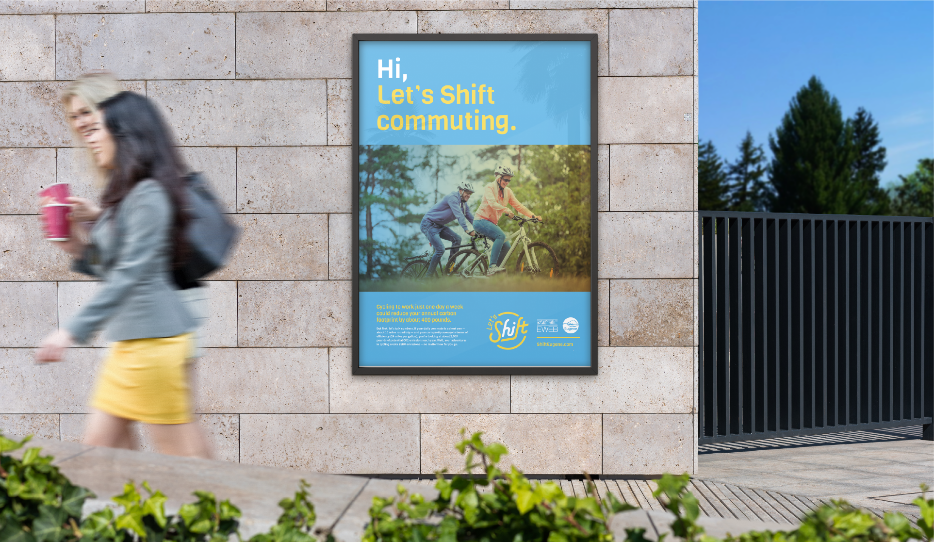 Framed 'Hi, Let's Shift commuting' bike poster mounted on a stone wall as pedestrians pass
