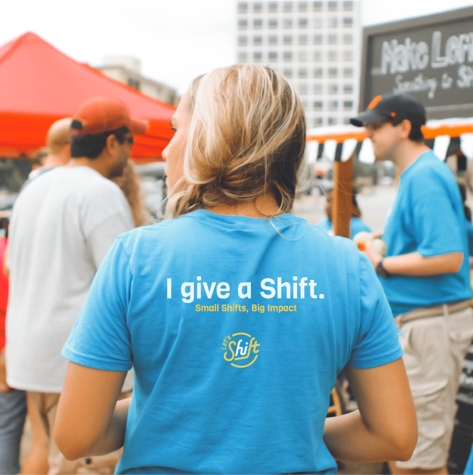 Woman seen from behind in a blue I give a Shift, Small Shifts Big Impact tee at an outdoor event