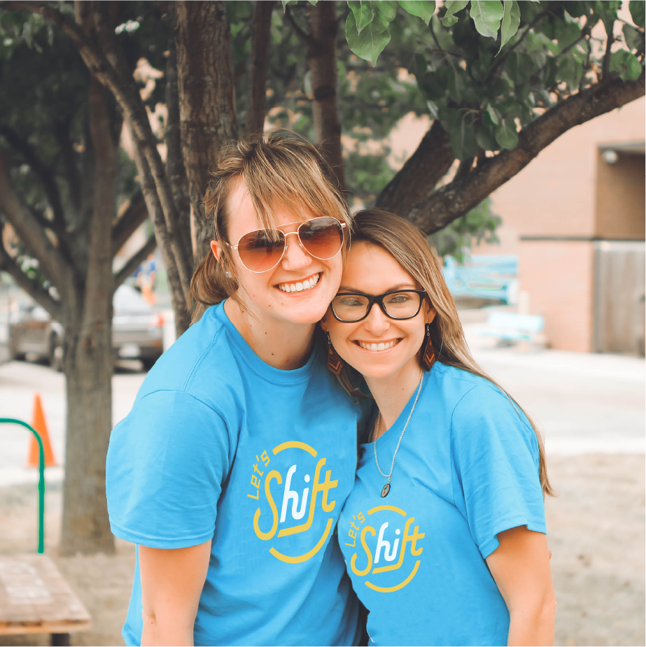 Two smiling women outdoors wearing blue "Let's Shift" t-shirts with yellow circular logo