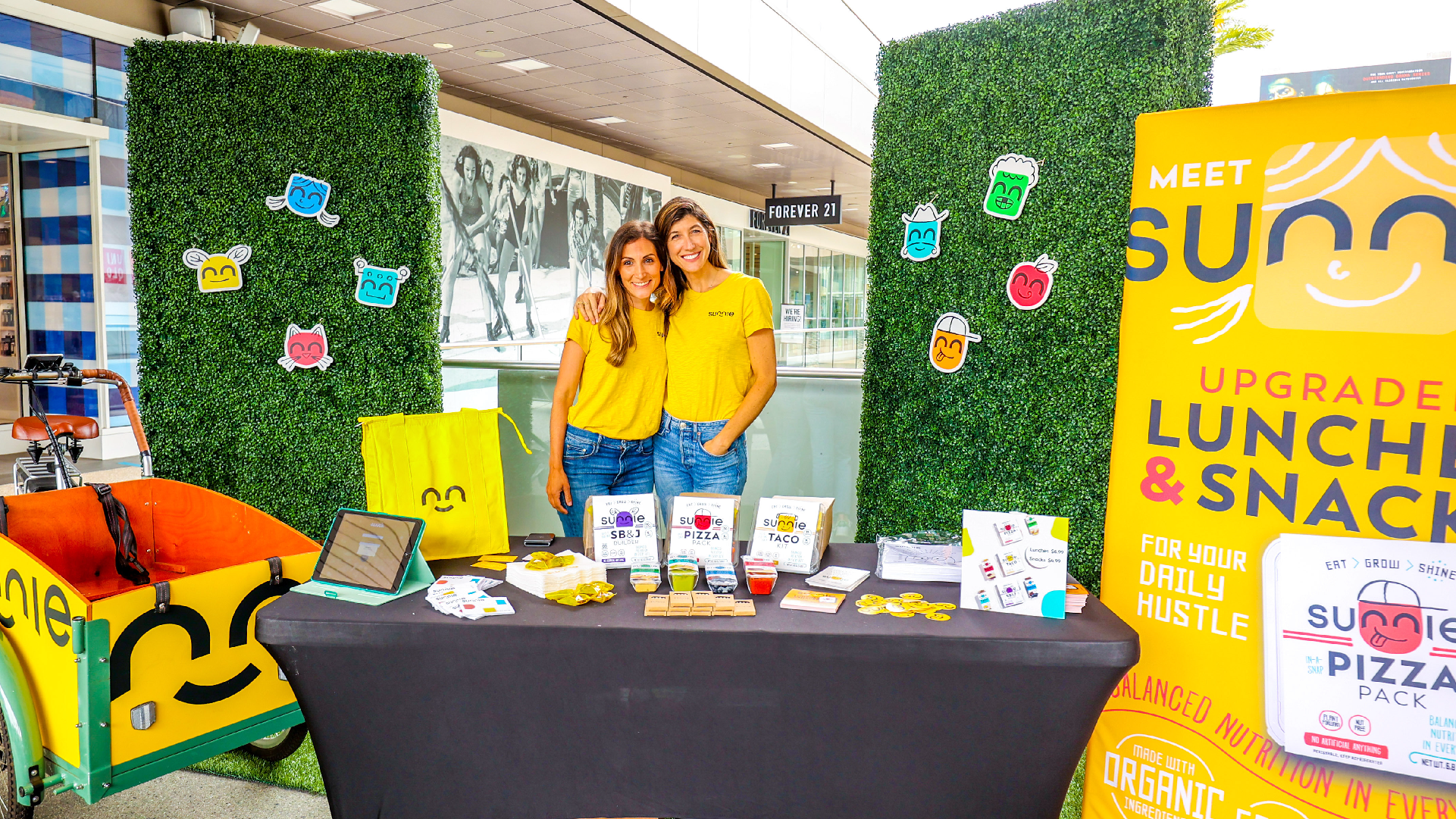 Two women in yellow Sunnie shirts at a pop-up booth with the Pizza Pack display and 'Meet Sunnie' banner