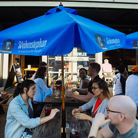 People drinking beer at outdoor tables under a blue Weihenstephan umbrella at the Tap and Growler