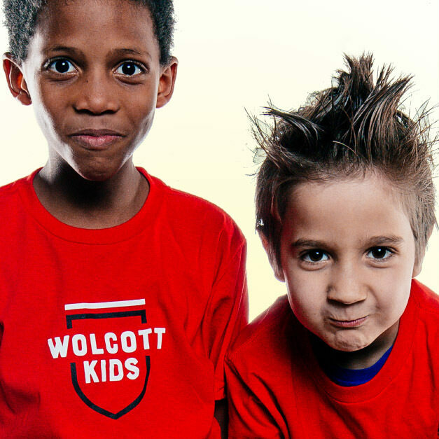 Two kids in red 'Wolcott Kids' shirts making playful faces, recruitment ad