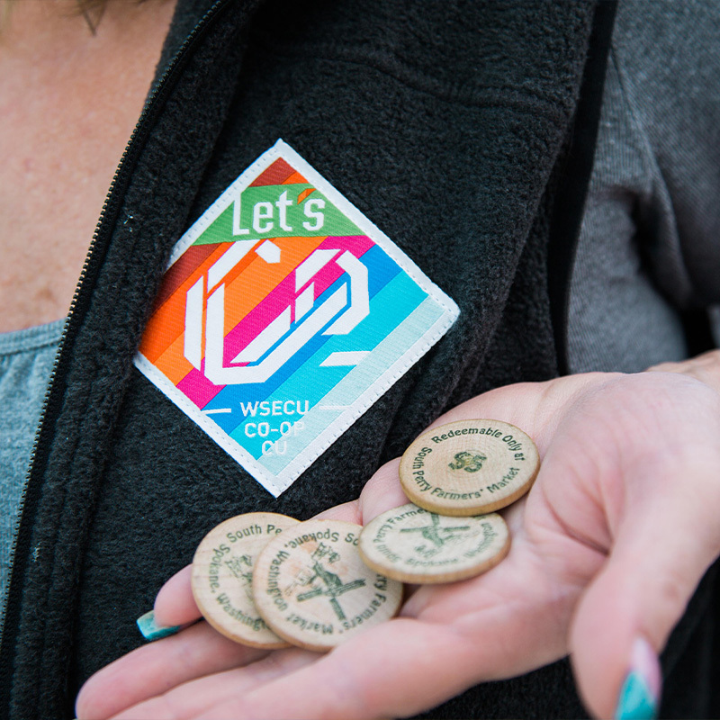 Colorful Let's Co WSECU co-op fabric badge pinned to a jacket beside wooden market tokens in a hand