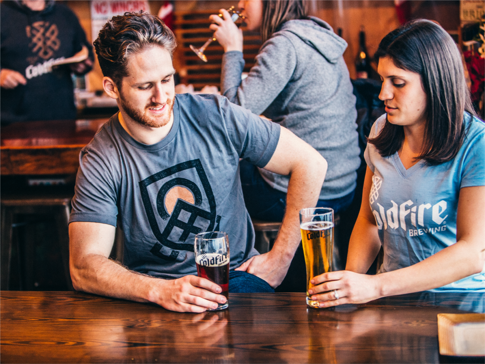 Couple in ColdFire Brewing shirts sharing pints of beer at a wood bar in the taproom
