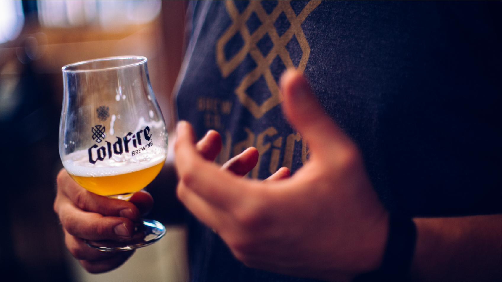 Hand holding a ColdFire Brewing tulip glass of beer beside a shirt with the embroidered hop logo