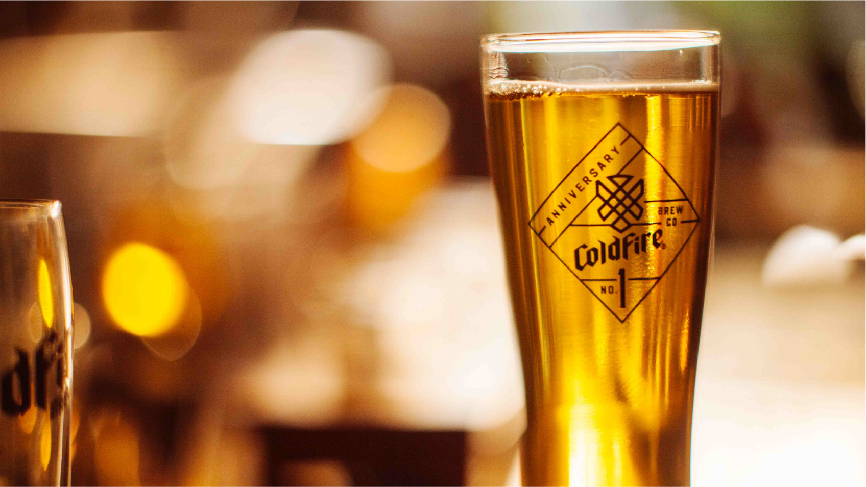 Tall glass of golden ColdFire beer with the anniversary diamond logo, warm bokeh lights behind