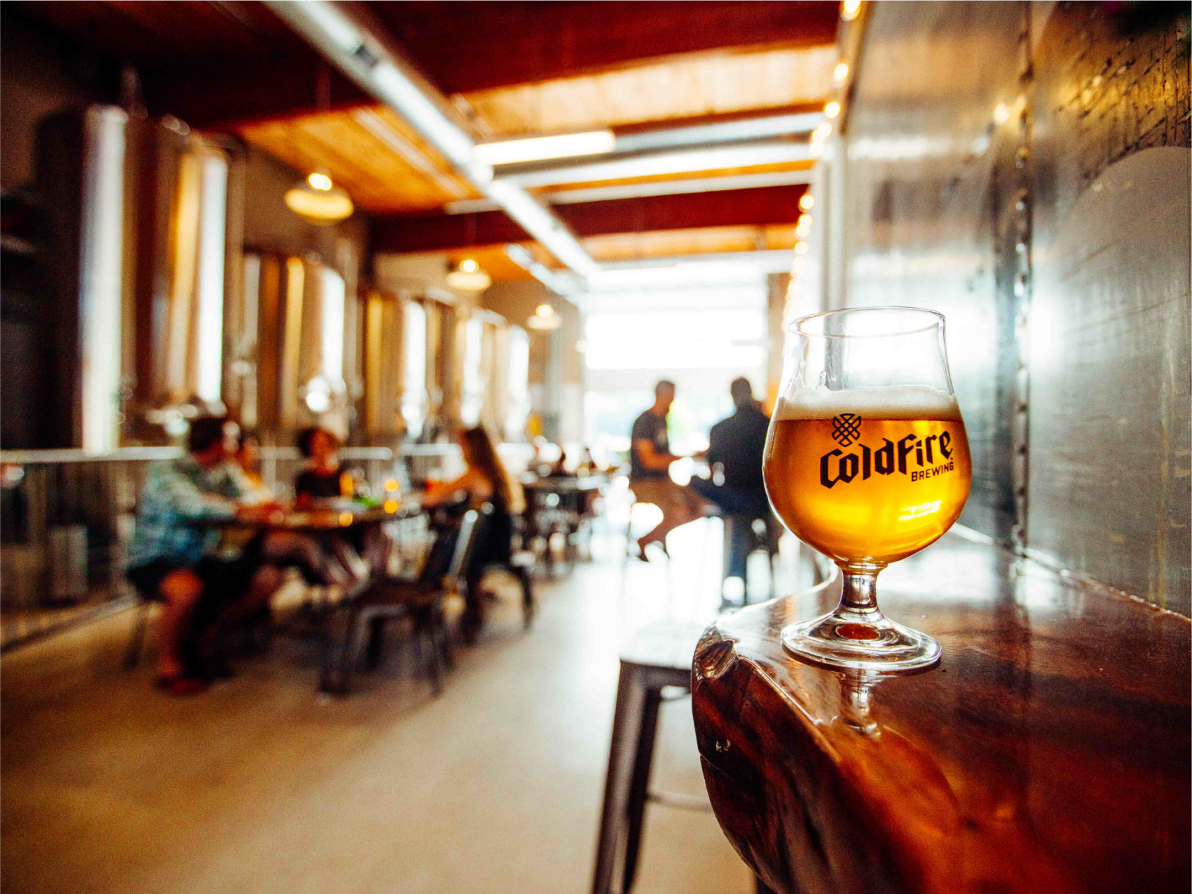 Glass of ColdFire beer on a wooden ledge with the brewery taproom and fermentation tanks blurred behind