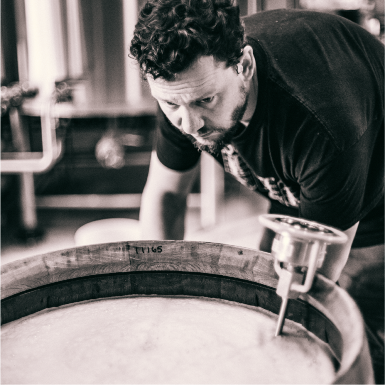 ColdFire brewer leaning over an oak barrel inspecting it, black and white