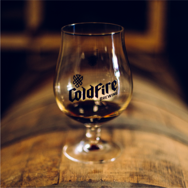 Snifter glass etched with the ColdFire Brewing logo holding beer atop a wooden barrel