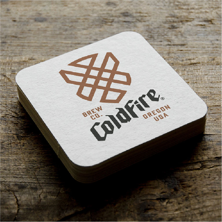 Stack of square ColdFire Brewing letterpress coasters with copper hop emblem on a weathered wood table
