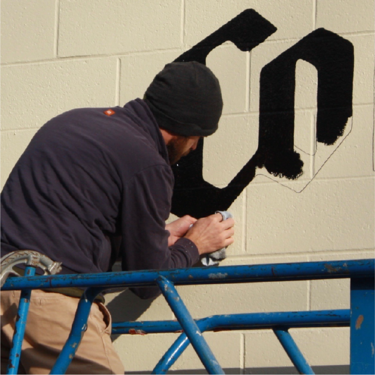Artist on a lift hand-painting the large black ColdFire Brewing logo on a cinder block wall