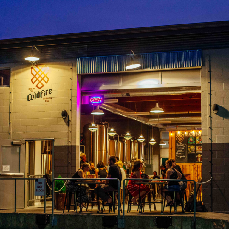 ColdFire Brewing taproom exterior at dusk with patrons seated outside and a neon Open sign
