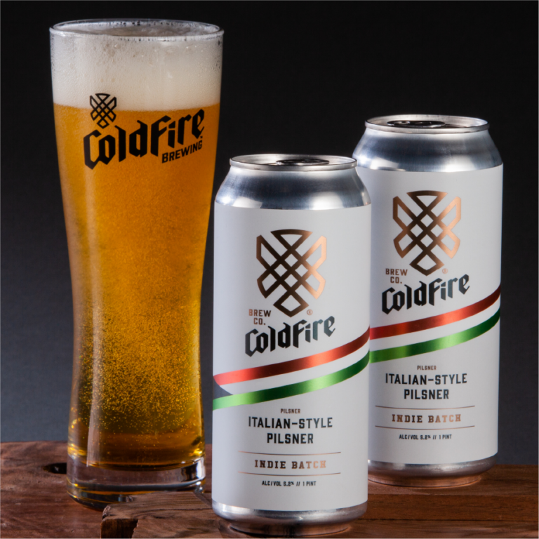ColdFire Brewing pilsner glass beside two cans of Italian-Style Pilsner Indie Batch on dark wood