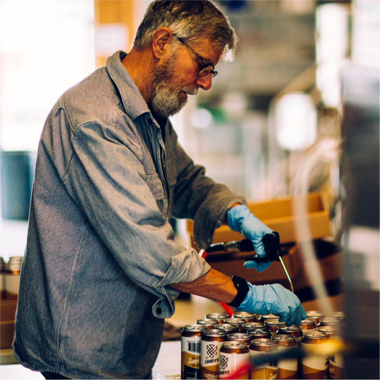 Bearded ColdFire worker in blue gloves hand-packing cans of beer in the brewery
