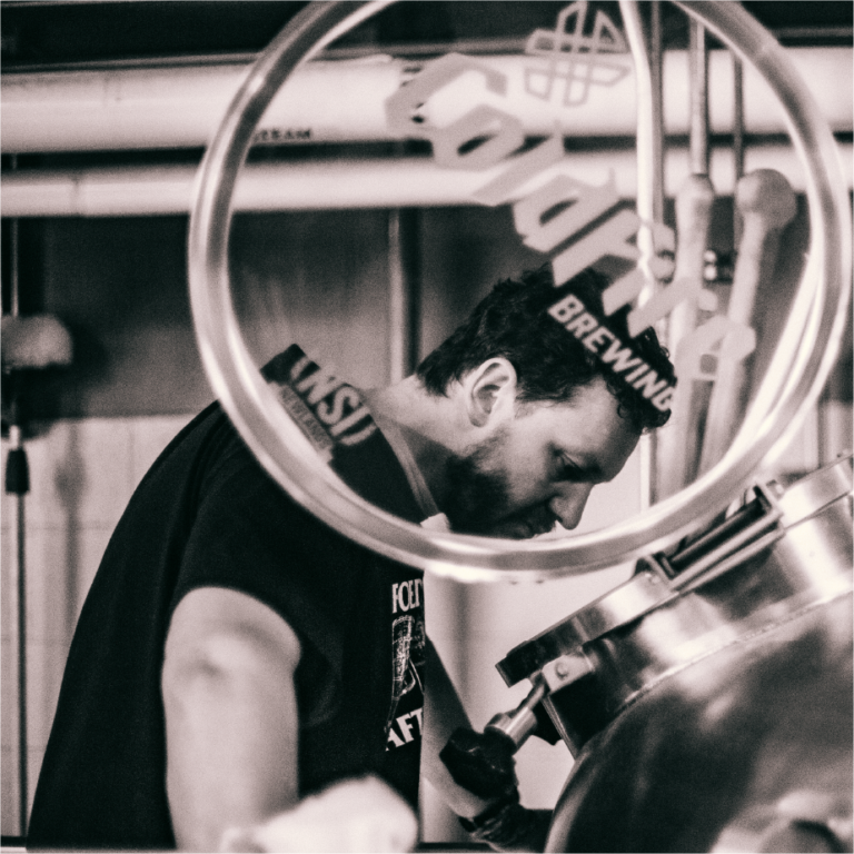 Black-and-white shot of a ColdFire Brewing worker bent over a steel tank with a process line overhead