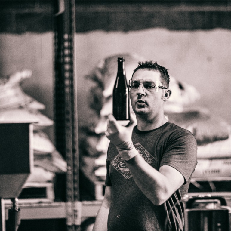Black-and-white shot of a ColdFire brewer in safety glasses holding up a bottle to inspect it