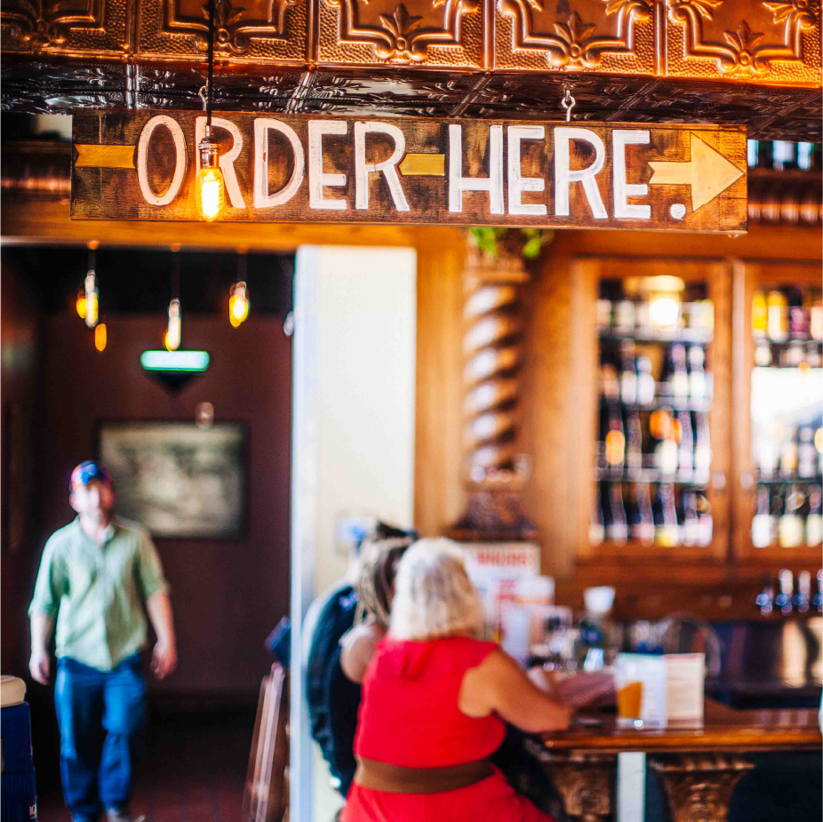 Pub interior with a hanging wooden "Order Here" arrow sign and bottle-lined bar