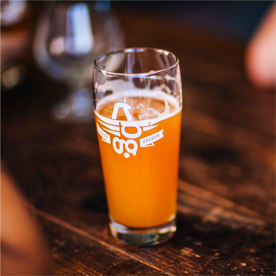 Glass of hazy orange beer with a winged "68" Oregon logo on a wood table