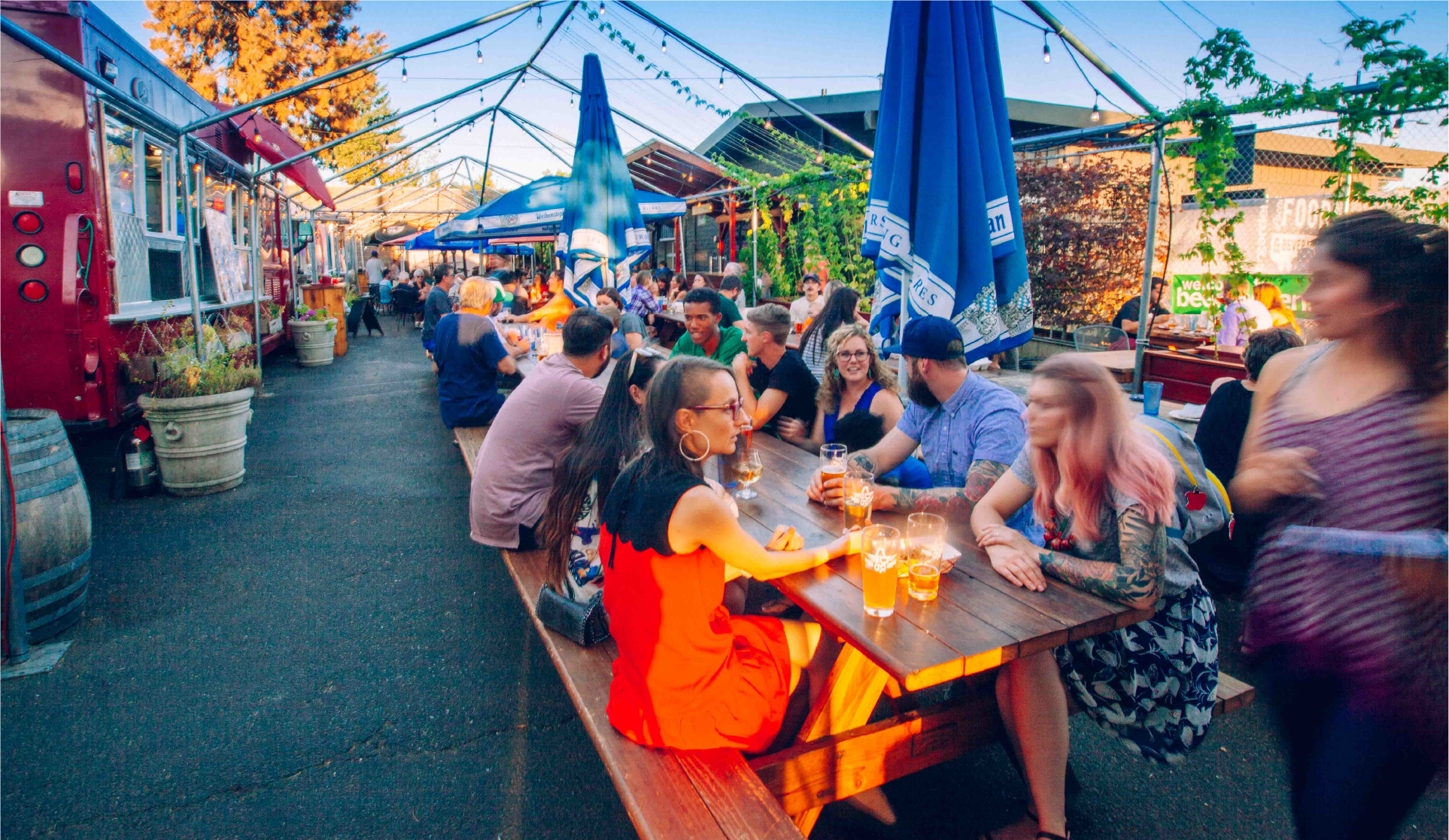 Crowd at outdoor beer garden picnic tables with food trucks and blue umbrellas at dusk