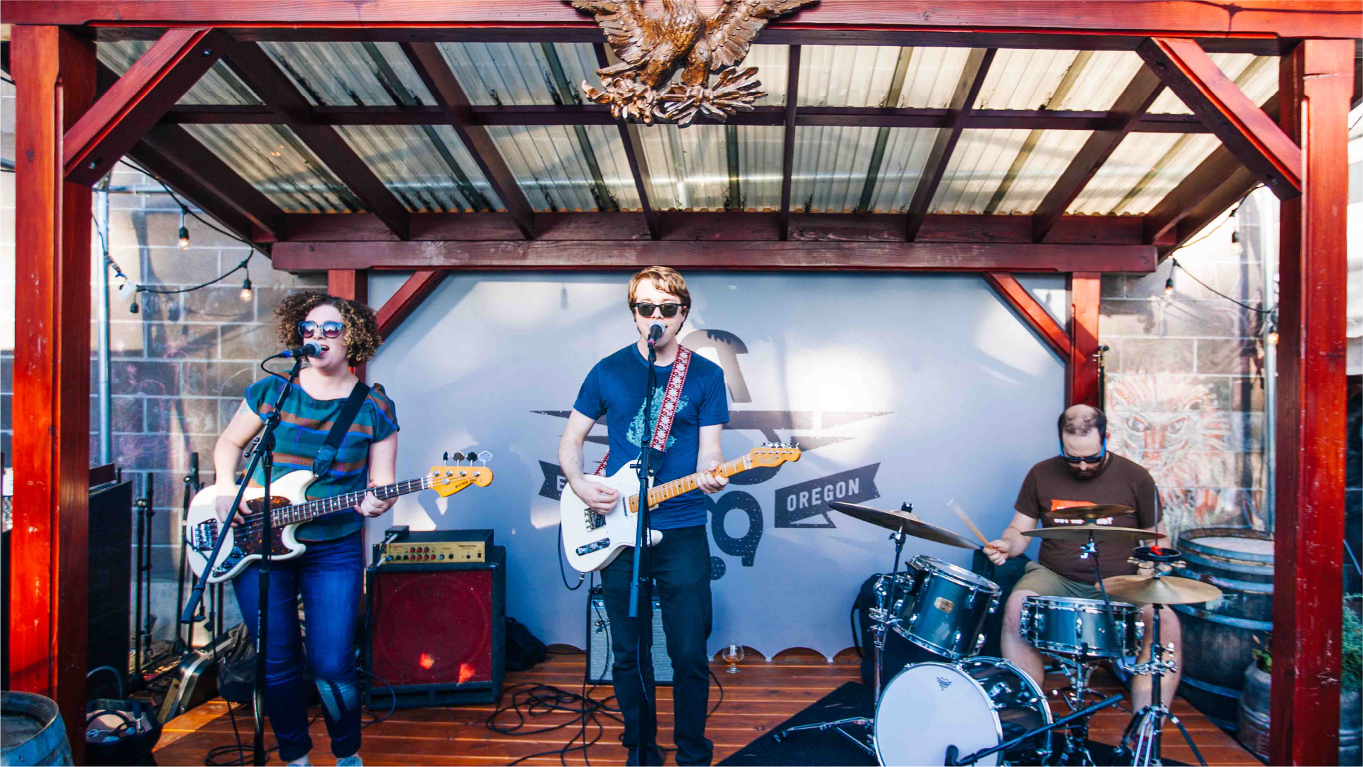 Band performing under a covered patio stage, guitarists and drummer at an outdoor venue