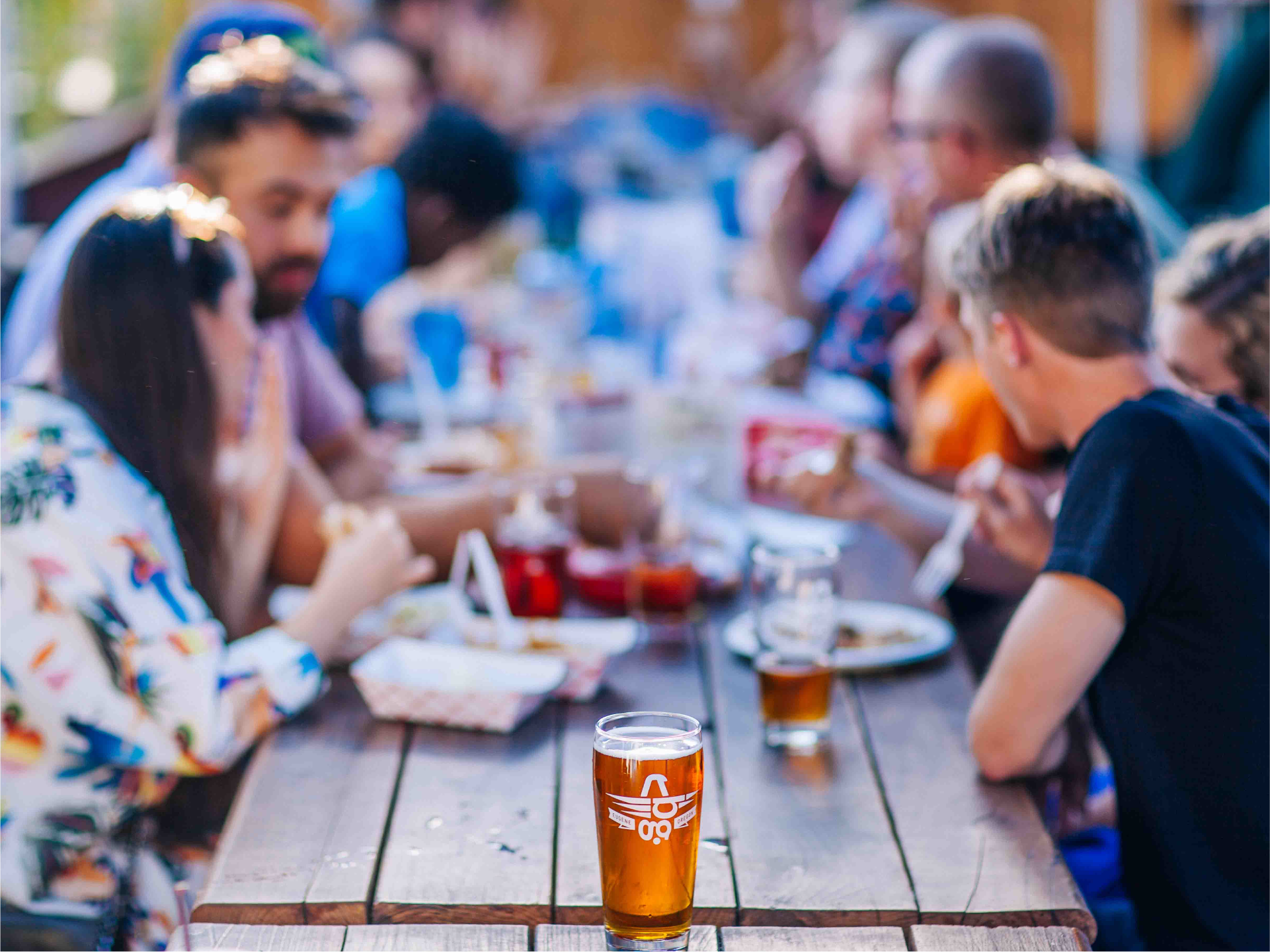 People dining at a long outdoor table with a beer glass etched with a logo in the foreground