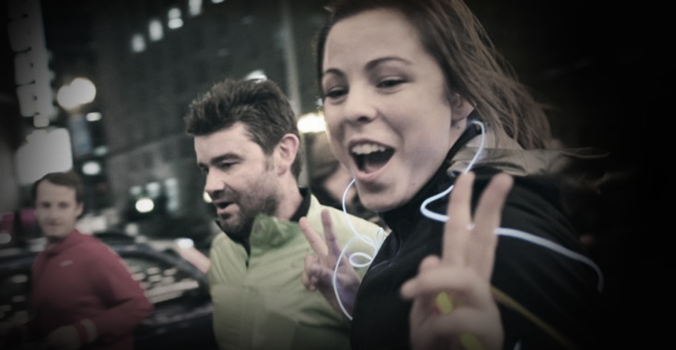 Two night runners with earphones smiling and flashing peace signs in a city, Nike London