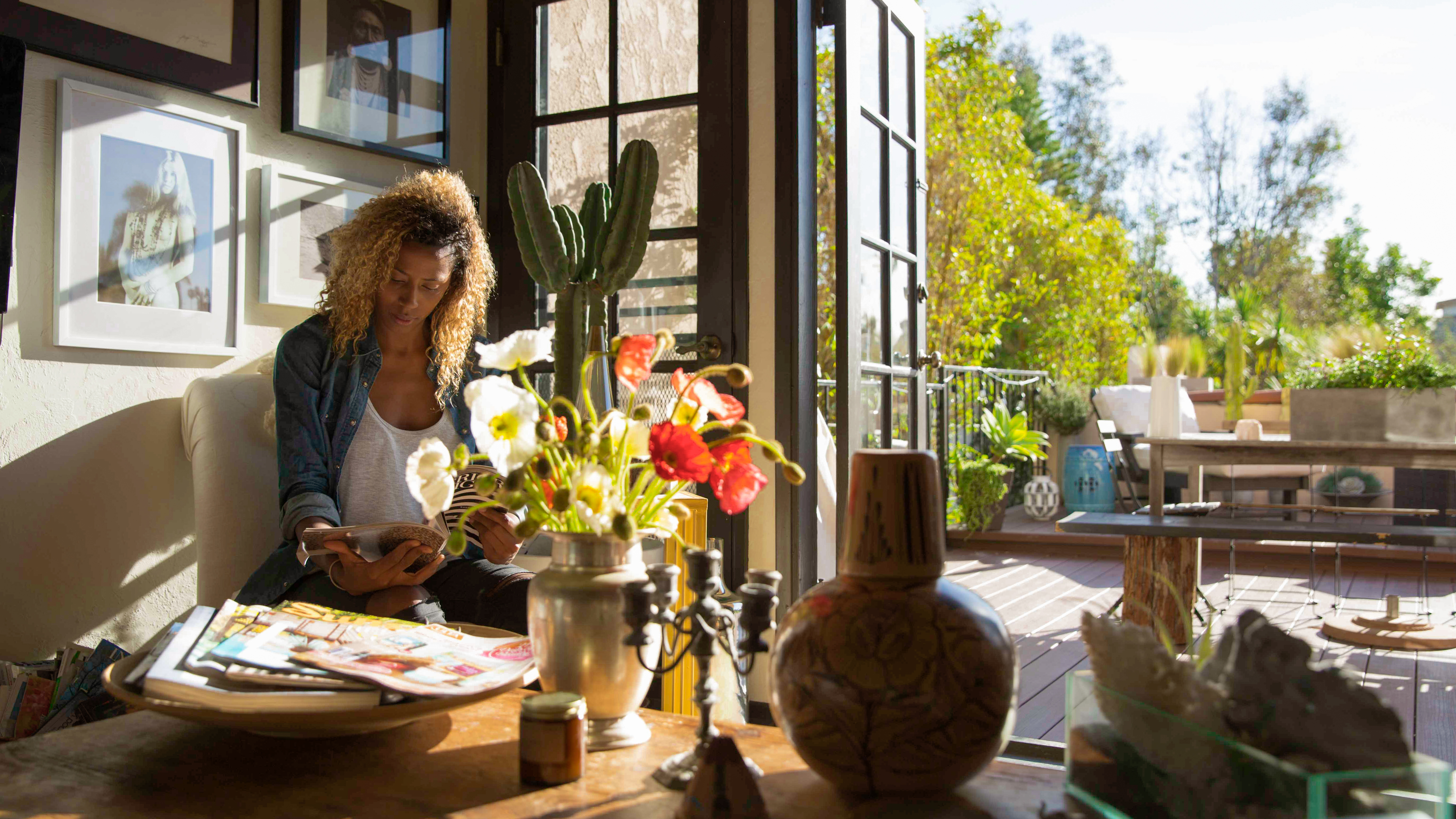 Woman reading a magazine by sunlit windows in a styled living room with flowers and pottery
