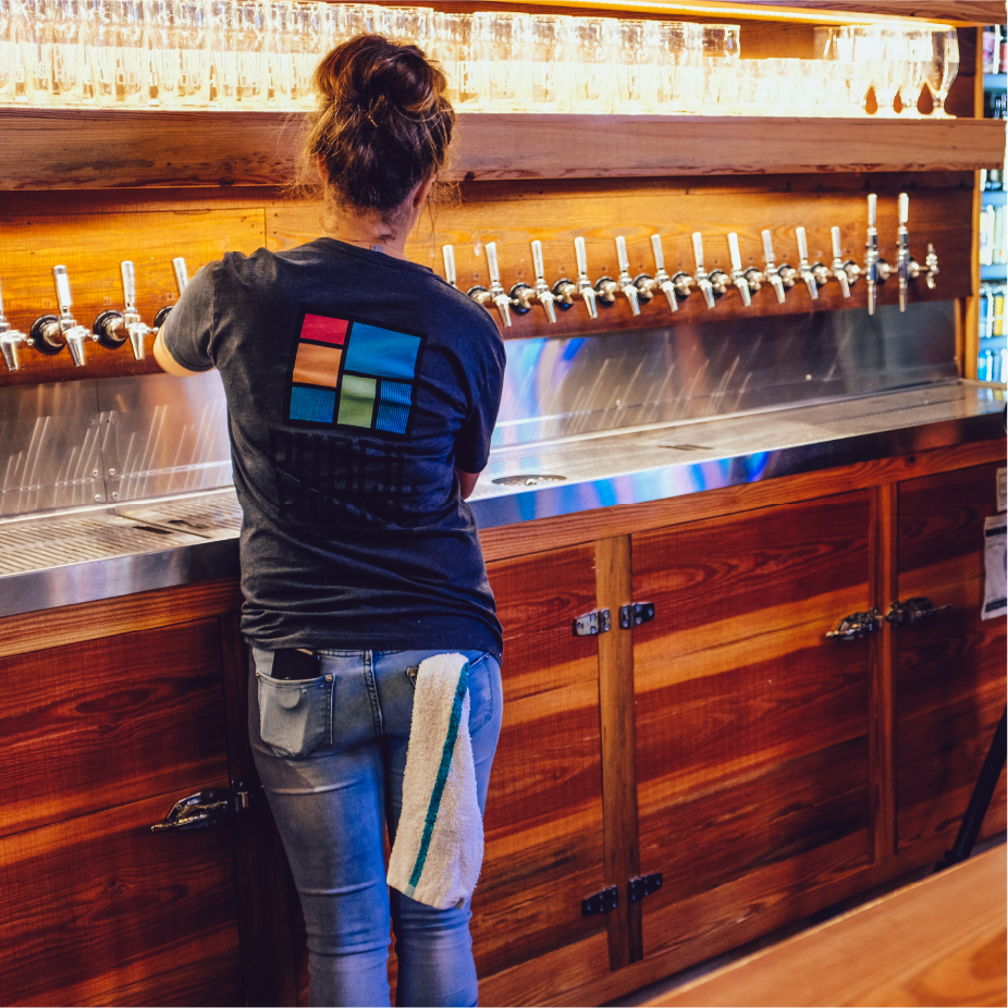 Bartender in Public House logo tee pouring from a long row of taps at a wooden bar