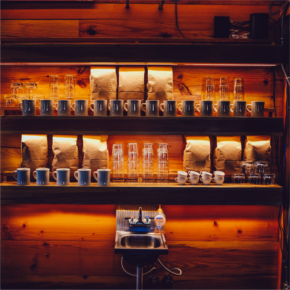 Backlit wood shelves of a coffee bar holding mugs, glasses and bagged coffee above a small sink