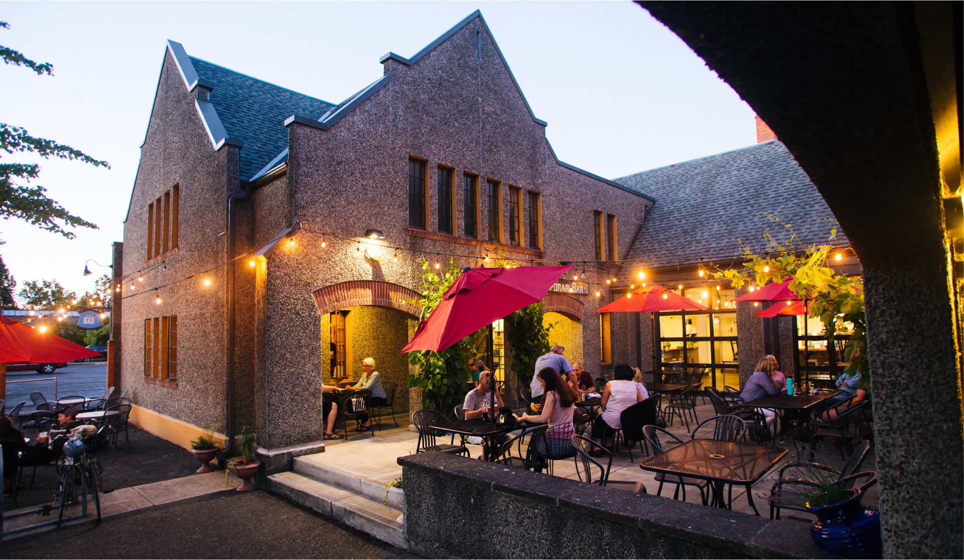 Stone restaurant building at dusk with patio diners under red umbrellas and string lights
