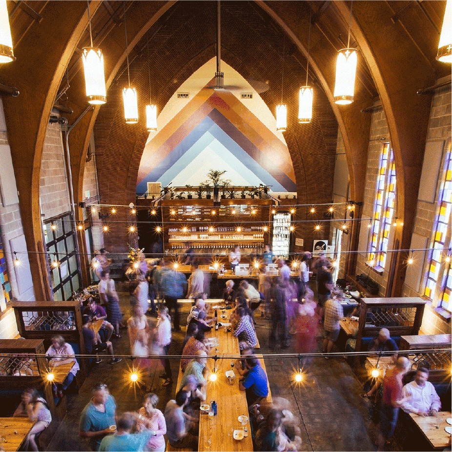 Crowded brewery in a converted church with arched ceilings, string lights and stained glass windows