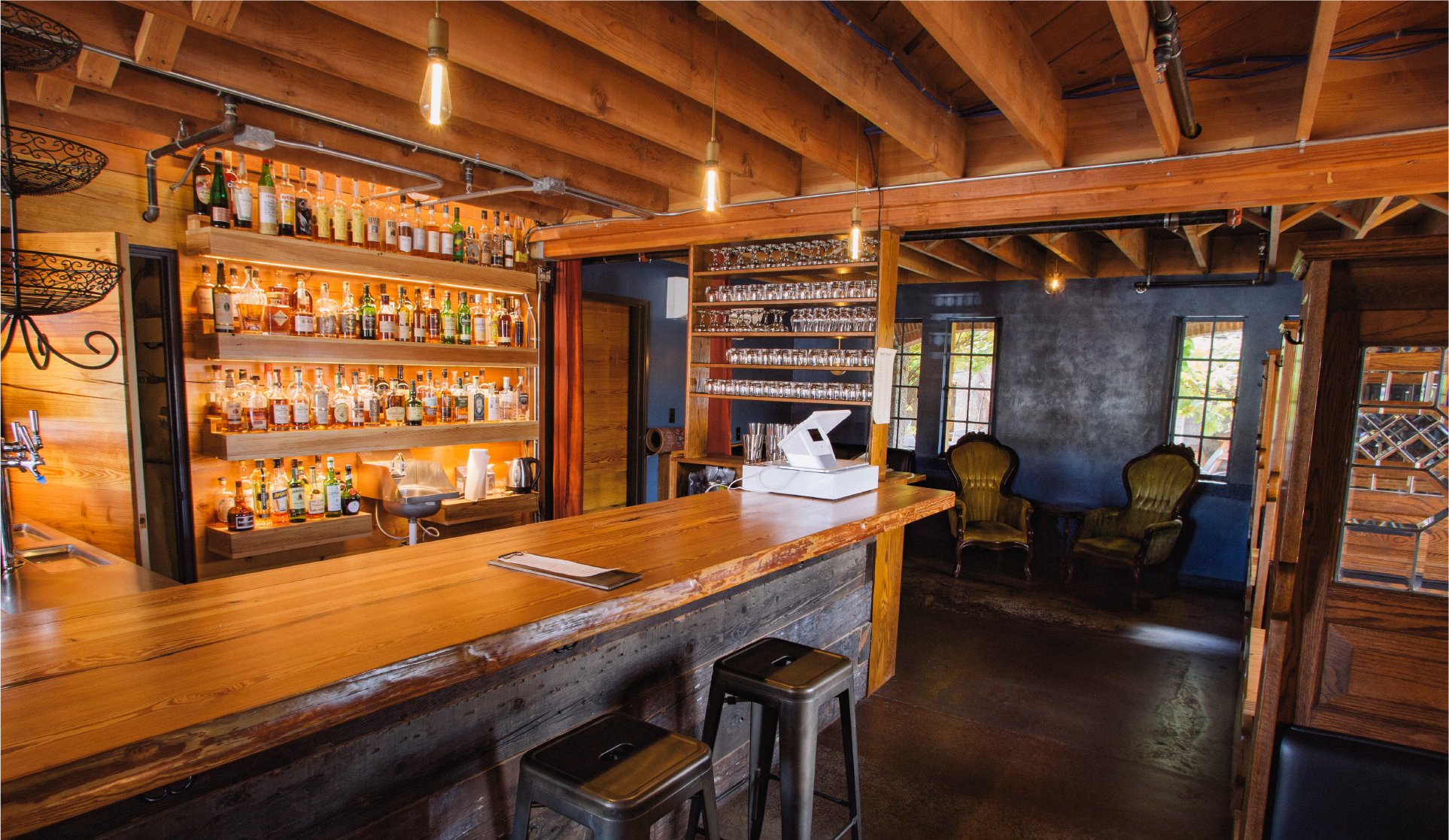 Public house interior with long wood bar, hanging Edison bulbs, and lounge chairs by windows