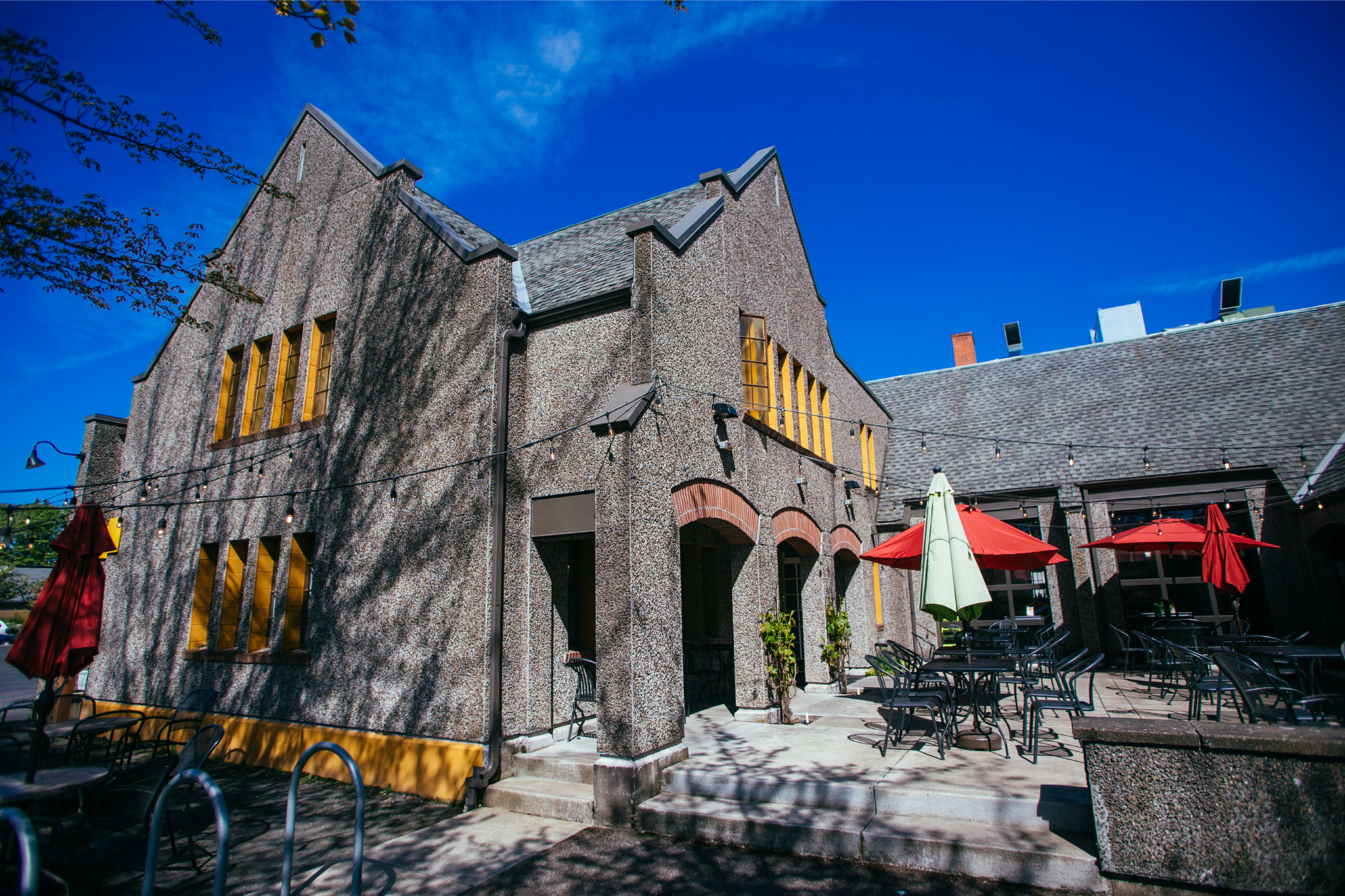 Stone Tudor-style building exterior with yellow trim and a patio with red umbrellas under a blue sky