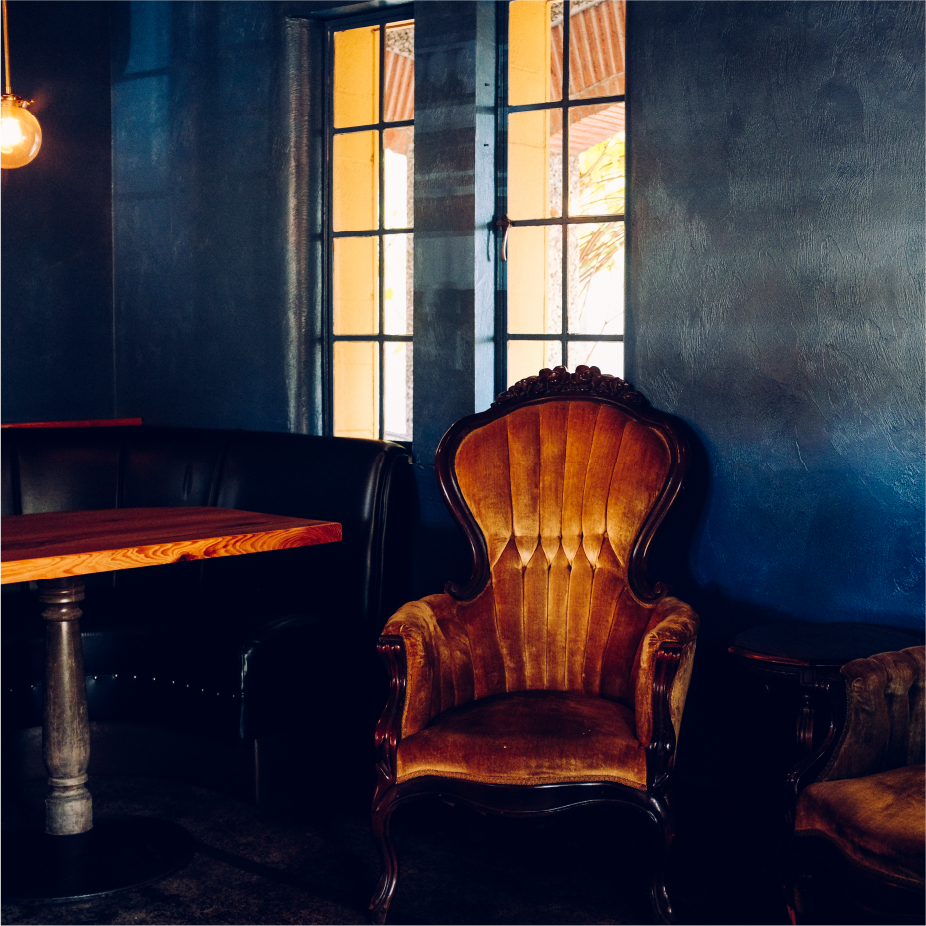 Dim bar interior with a velvet orange armchair, wood table, and tall windows with warm light