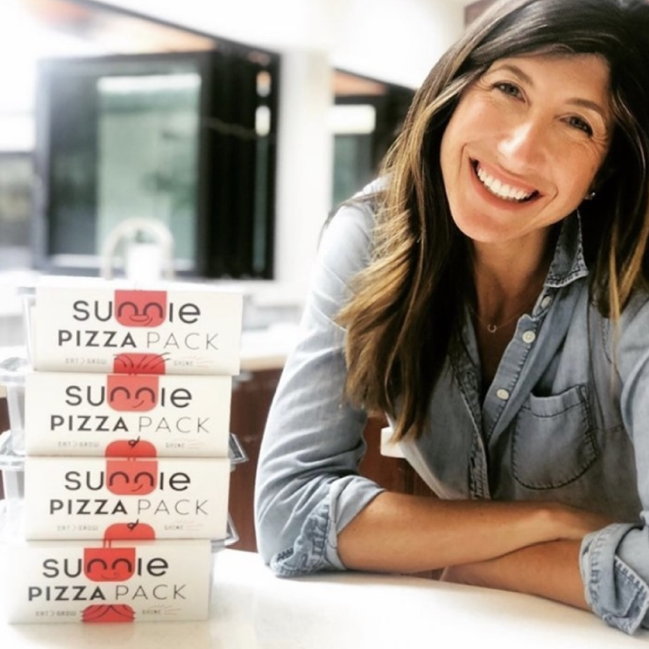 Smiling woman leaning on a counter beside a stack of four Sunnie Pizza Pack lunch boxes