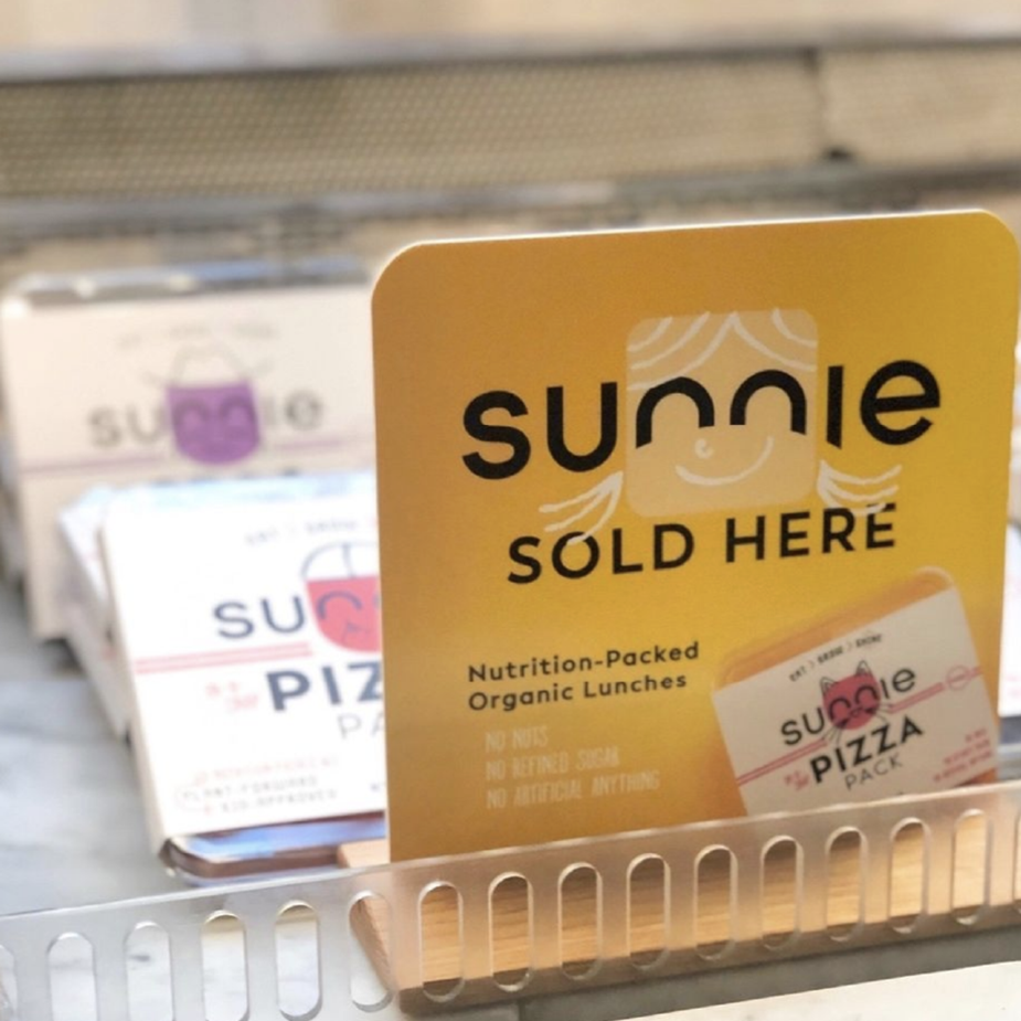 Retail shelf sign reading 'Sunnie sold here, nutrition-packed organic lunches' above Sunnie Pizza Pack boxes