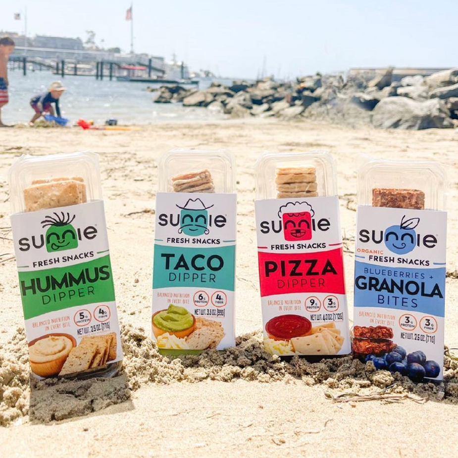 Sunnie Fresh Snacks Hummus, Taco, Pizza and Granola dipper packs standing in beach sand by the water
