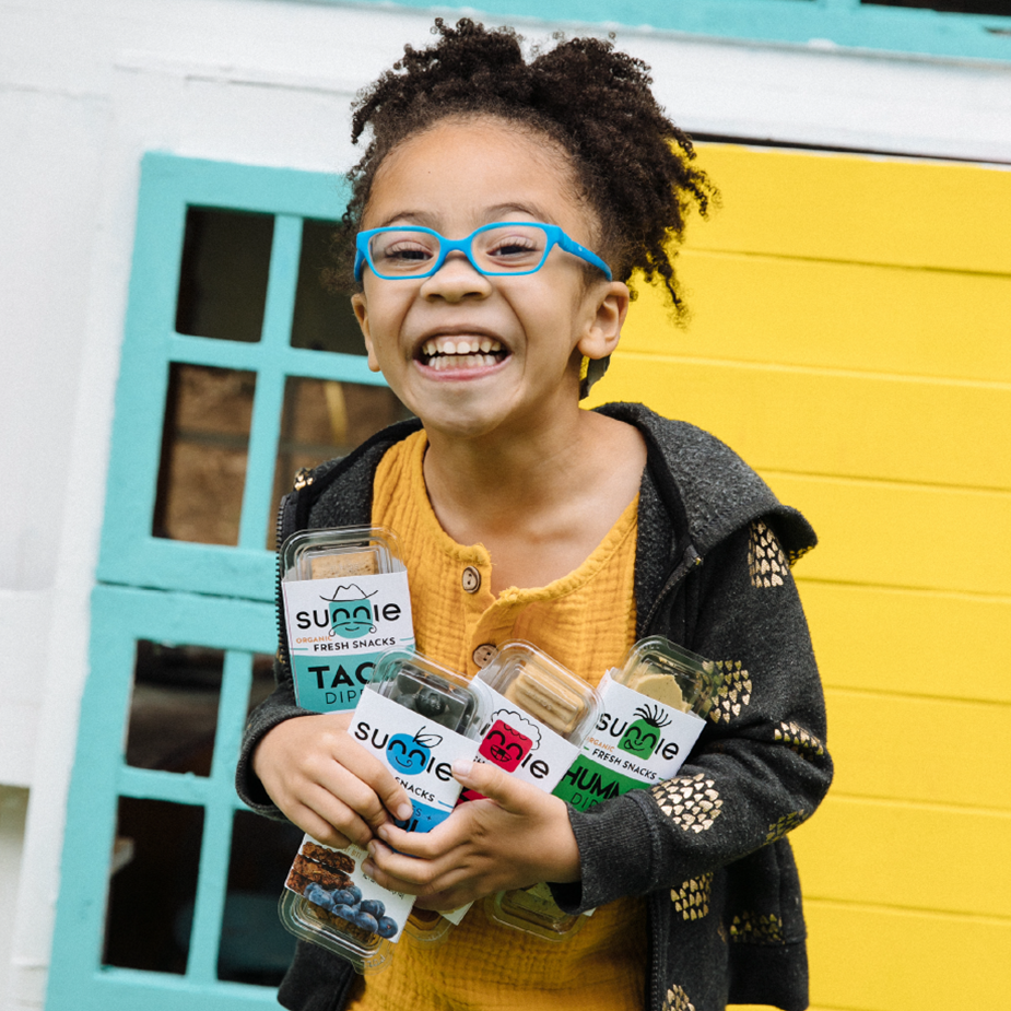 Smiling child in blue glasses holds an armful of Sunnie Fresh Snacks dipper packs outside a playhouse