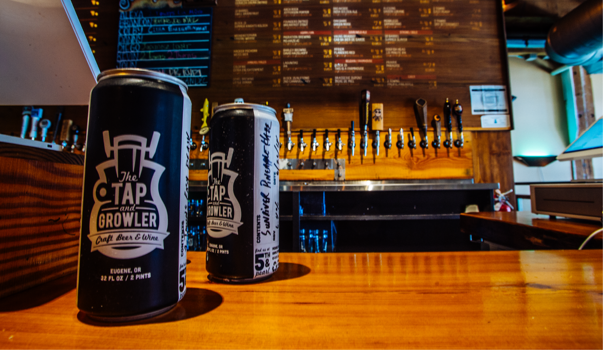 Two black Tap and Growler crowler cans on a bar counter with taps and menu board behind